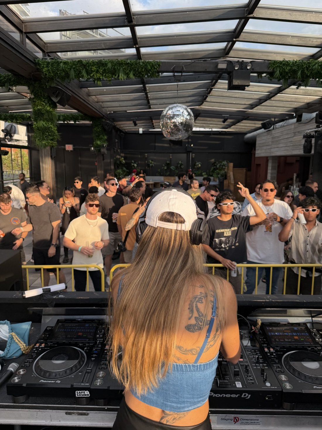 Sierra Bomba behind the decks facing a crowd beneath a disco ball and glass roof
